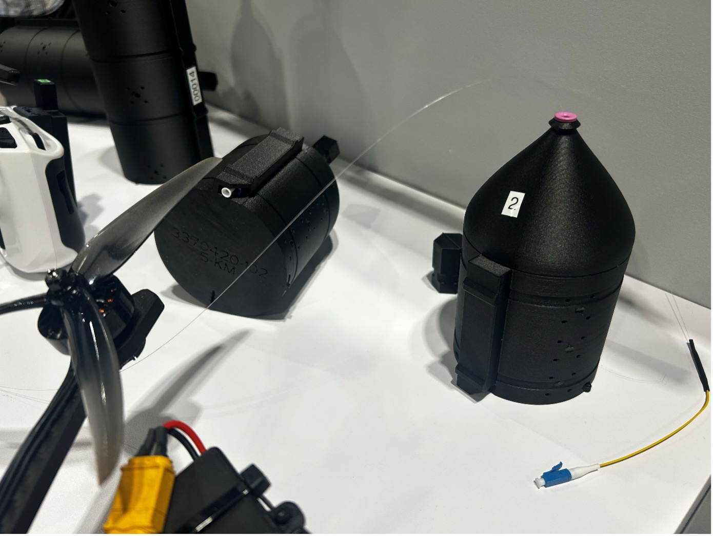 Die Gehäuse mit dem Glasfaserkabel – die schwarzen Behälter in verschiedenen Größen – werden unter der Drohne angebracht und wickeln sich während des Fluges ab. Der blaue Stecker kommt als Verbindung in den Kontroller.